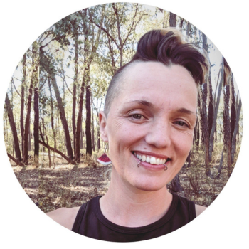 Smiling person with short styled hair standing in a sunlit forest setting.